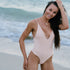 Model at a beach having reversed light pink Emerald Beach One Piece - Deep V plunging neckline Moderate back coverage Mid to high waist cut Adjustable straps, smiling and posing to set her hair